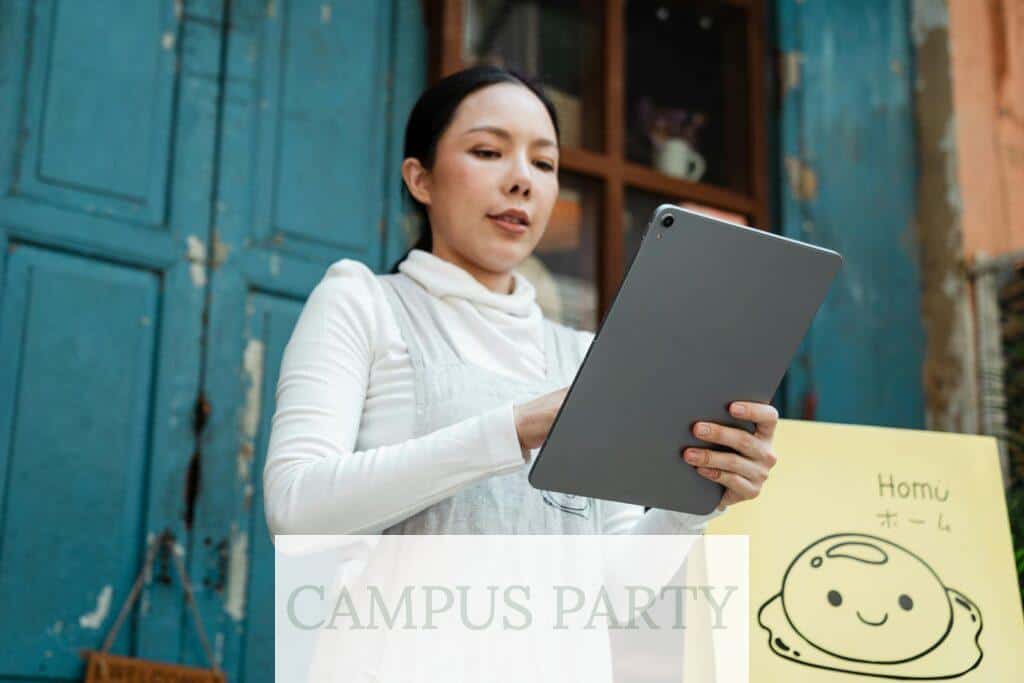 Positive Asian lady in white shirt and gray apron using modern tablet while standing near rural cafe door on daytime