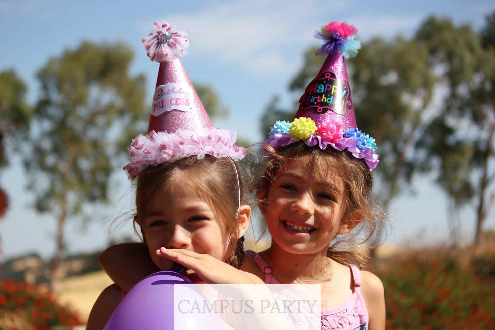 girl in left blowing purple balloon beside girl wearing pink birthday hat